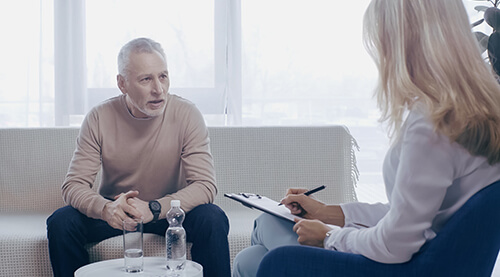 middle aged man talking with blonde psychologist holding pen and clipboard in consulting room